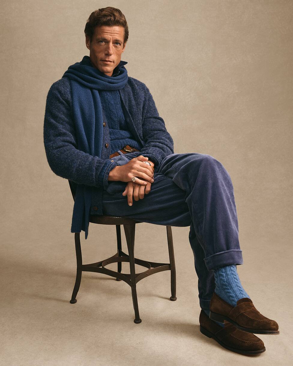 Homem sentado com camisola de malha azul e cachecol a condizer, com mocassins castanhos.