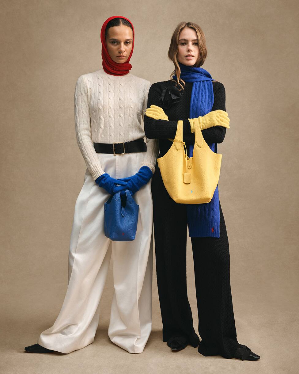 Duas mulheres de pé, uma com camisola de malha creme com gorro vermelho e luvas azuis, e a outra com camisola de malha preta com cachecol azul e mala amarela.