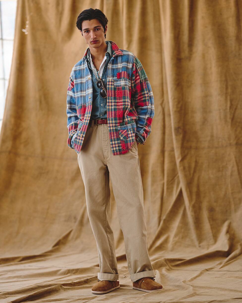 Homem com camisa de flanela sobre uma camisa de denim, com calças bege e mocassins.